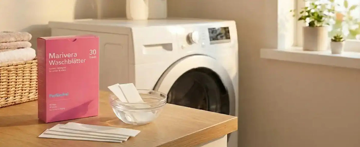 Eine rosa Packung „Marivera Waschblätter“ steht neben einer Schale mit weißen Waschmittelblätter auf einem Holztisch, im Hintergrund eine moderne weiße Waschmaschine in einem sonnendurchfluteten Raum.
